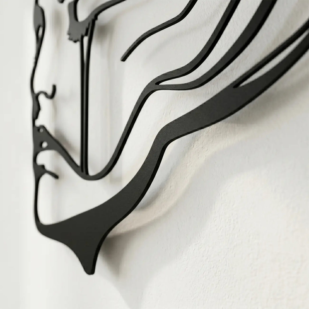 Detailopname van een zwarte metalen wanddecoratie (abstract gezicht) aan een lichte muur, die het 3D-effect en het dynamische schaduwspel door de afstandhouders benadrukt.